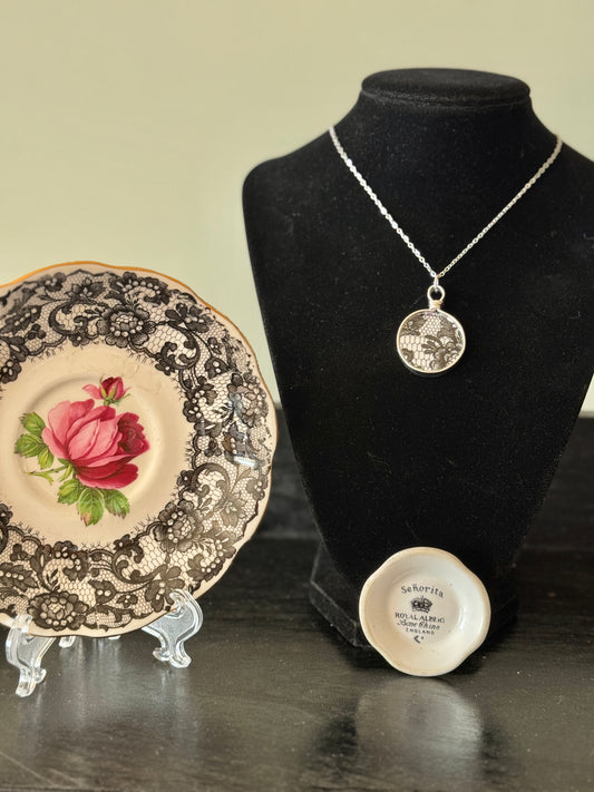 Necklace displayed on a black mannequin bust with a decorative plate and small round object on a reflective surface.