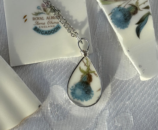 Teardrop-shaped pendant with floral design on a gray surface, with 'Royal Albert' branding in the background.