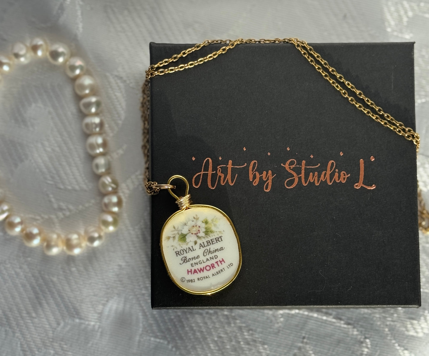 Necklace with a pendant on a black card labeled 'Art by Studio L' next to a pearl bracelet.