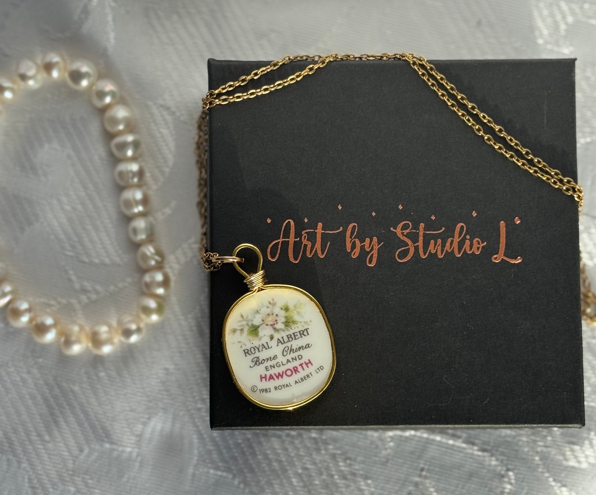 Necklace with a pendant on a black card labeled 'Art by Studio L' next to a pearl bracelet.