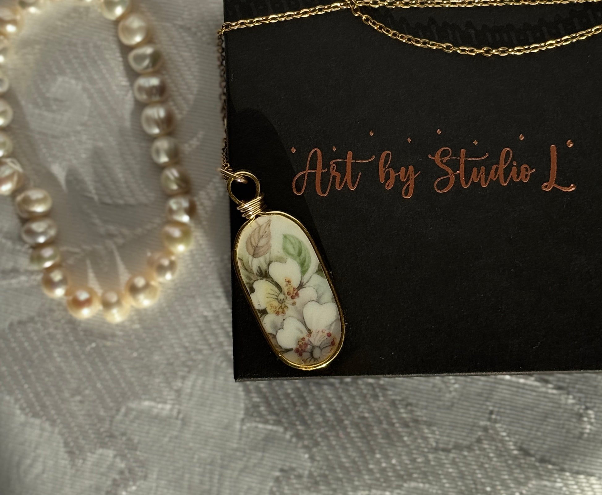 Necklace with a floral pendant on a black card with 'Art by Studio L' text, pearl necklace on gray fabric background