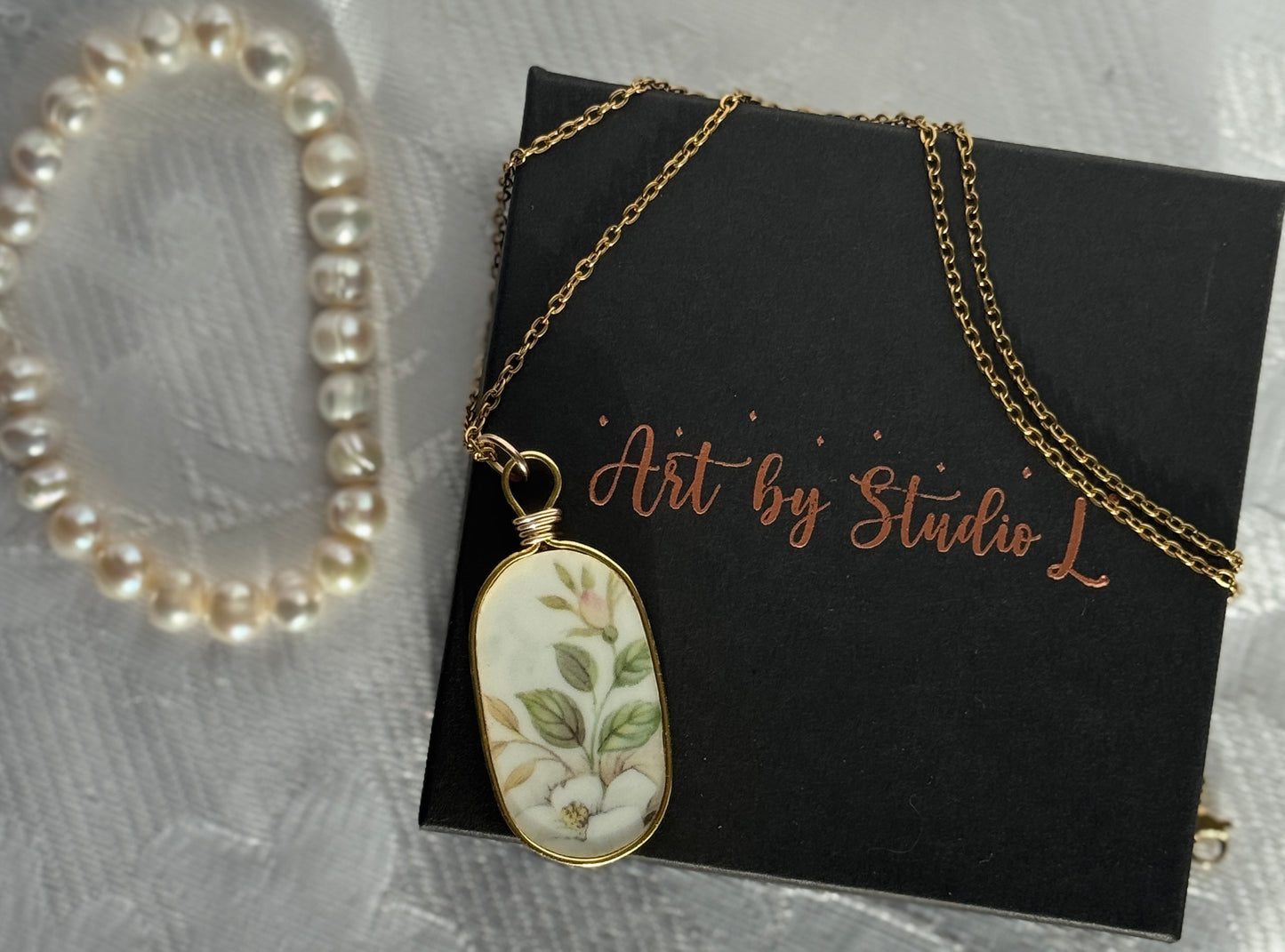 Necklace with a floral pendant on a black card with 'Art by Studio L' text, pearl bracelet in the background.