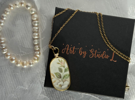 Necklace with a floral pendant on a black card with 'Art by Studio L' text, pearl bracelet in the background.