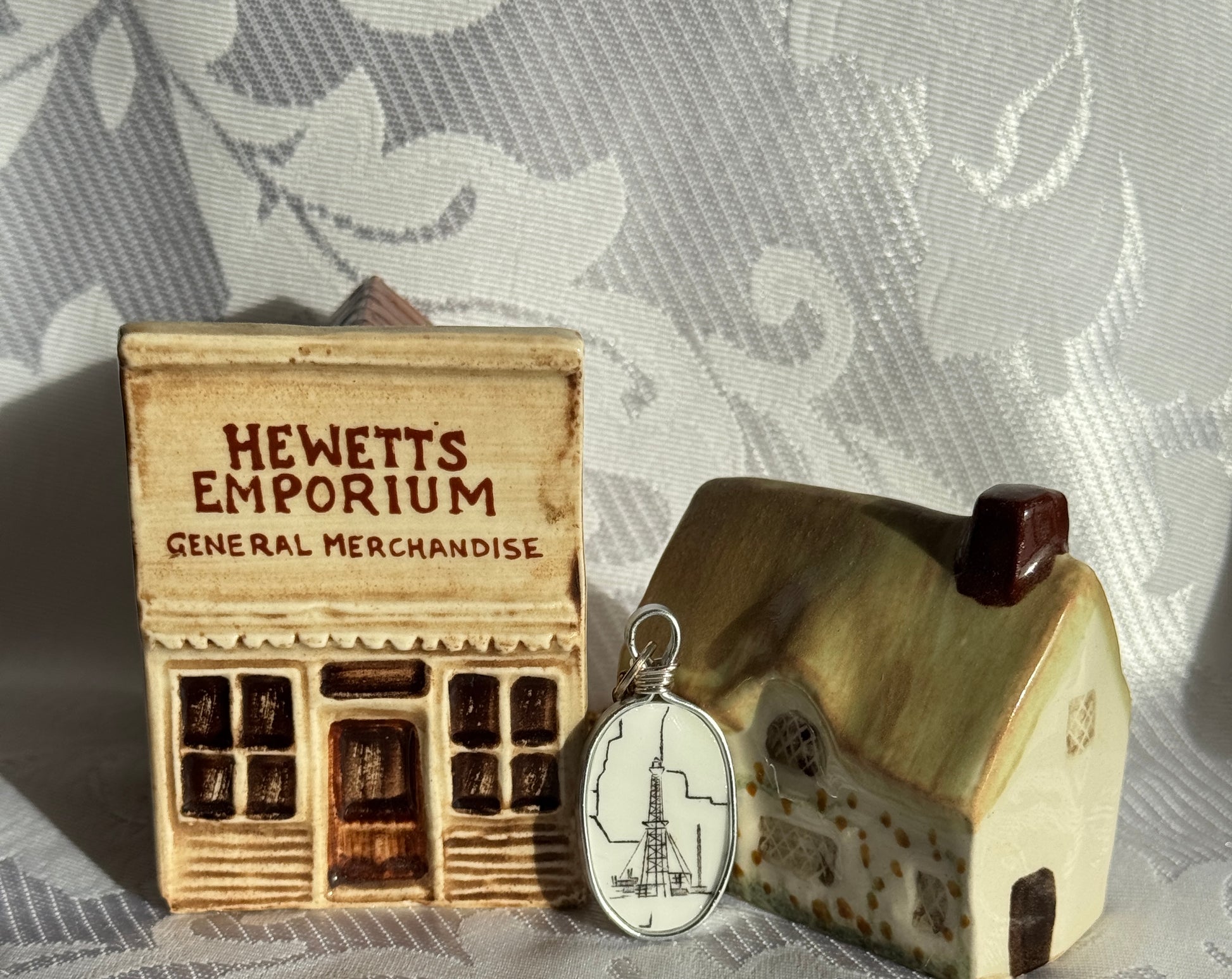 Small wooden model of a store with 'Hewetts Emporium' sign, next to a decorative house and a pendant on a textured surface.