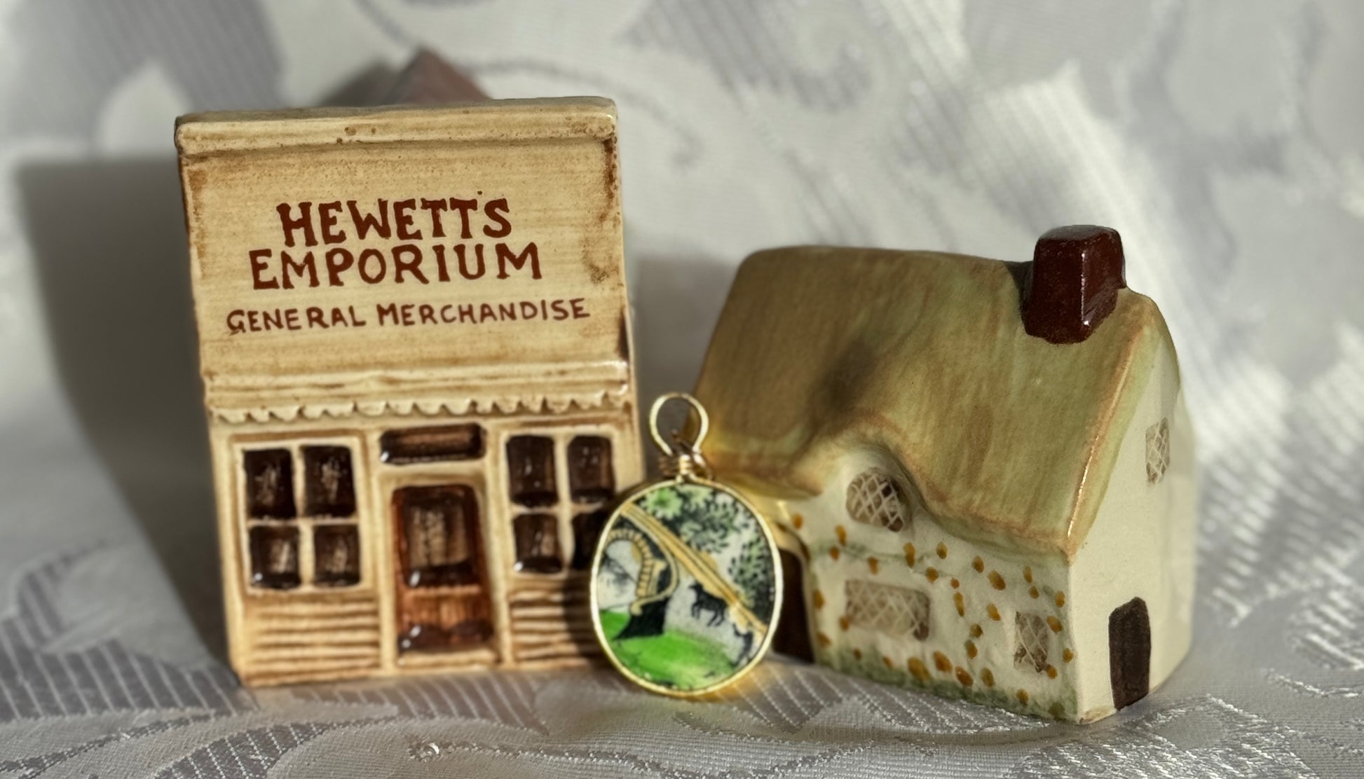 A pendant with a cow and a herder, beside miniature model of a store named 'Hewett's Emporium' and a house on a textured surface.
