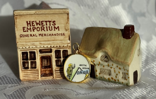 A pendant with a Inuit beside two small models of a building, a house, and a sign with 'Hewett's Emporium' on a textured surface.