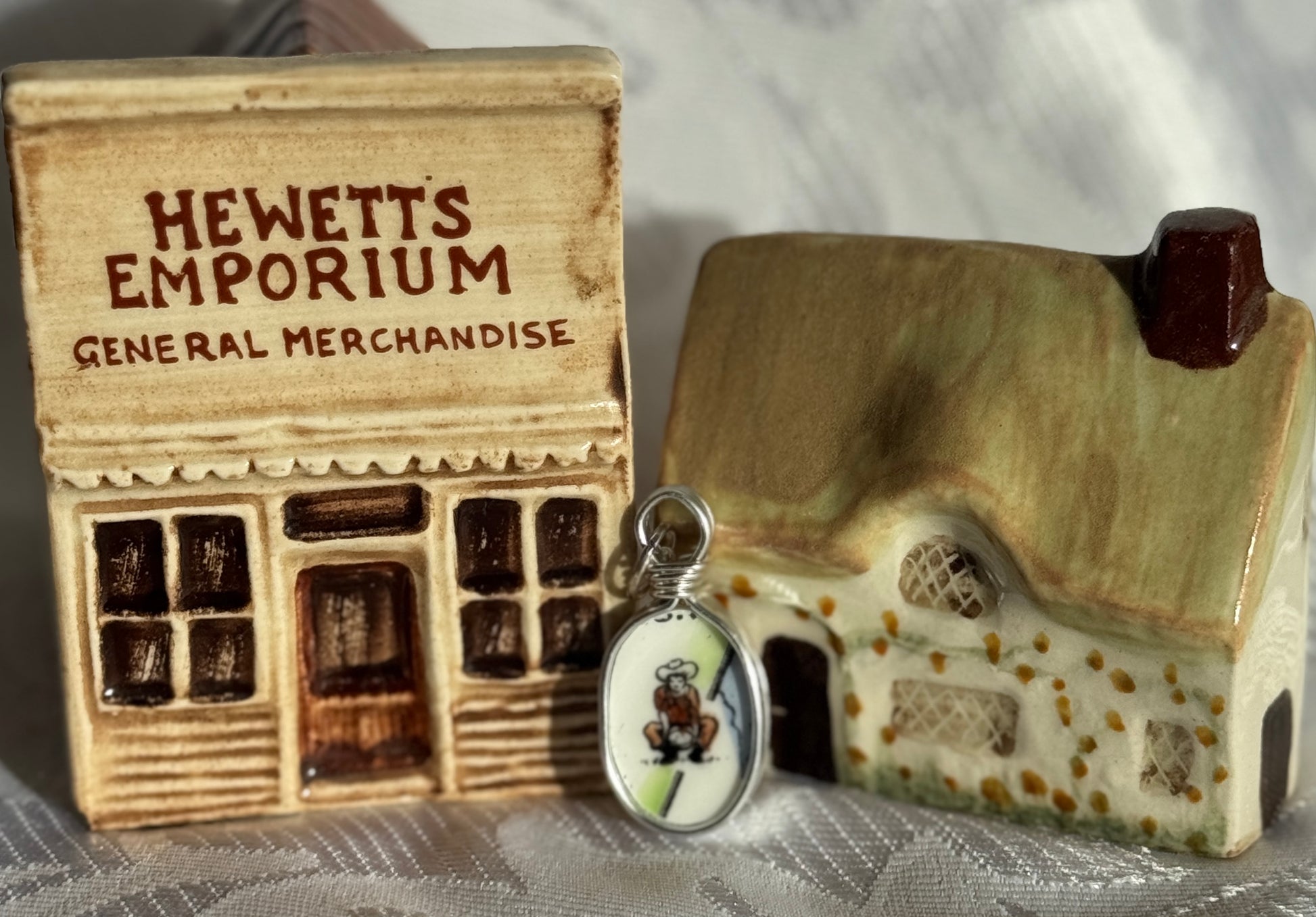 A cowboy on a pendant, beside a small model of a store front with 'Hewetts Emporium' and a miniature house on a textured surface.