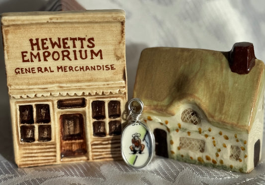 A cowboy on a pendant, beside a small model of a store front with 'Hewetts Emporium' and a miniature house on a textured surface.
