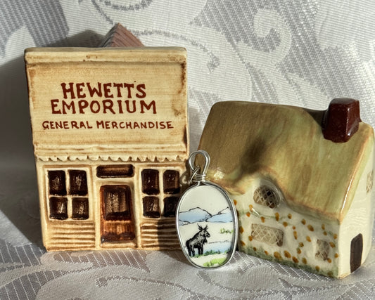 Small wooden model of a store with 'Hewett's Emporium' and a pendant with a Moose design on a textured surface.