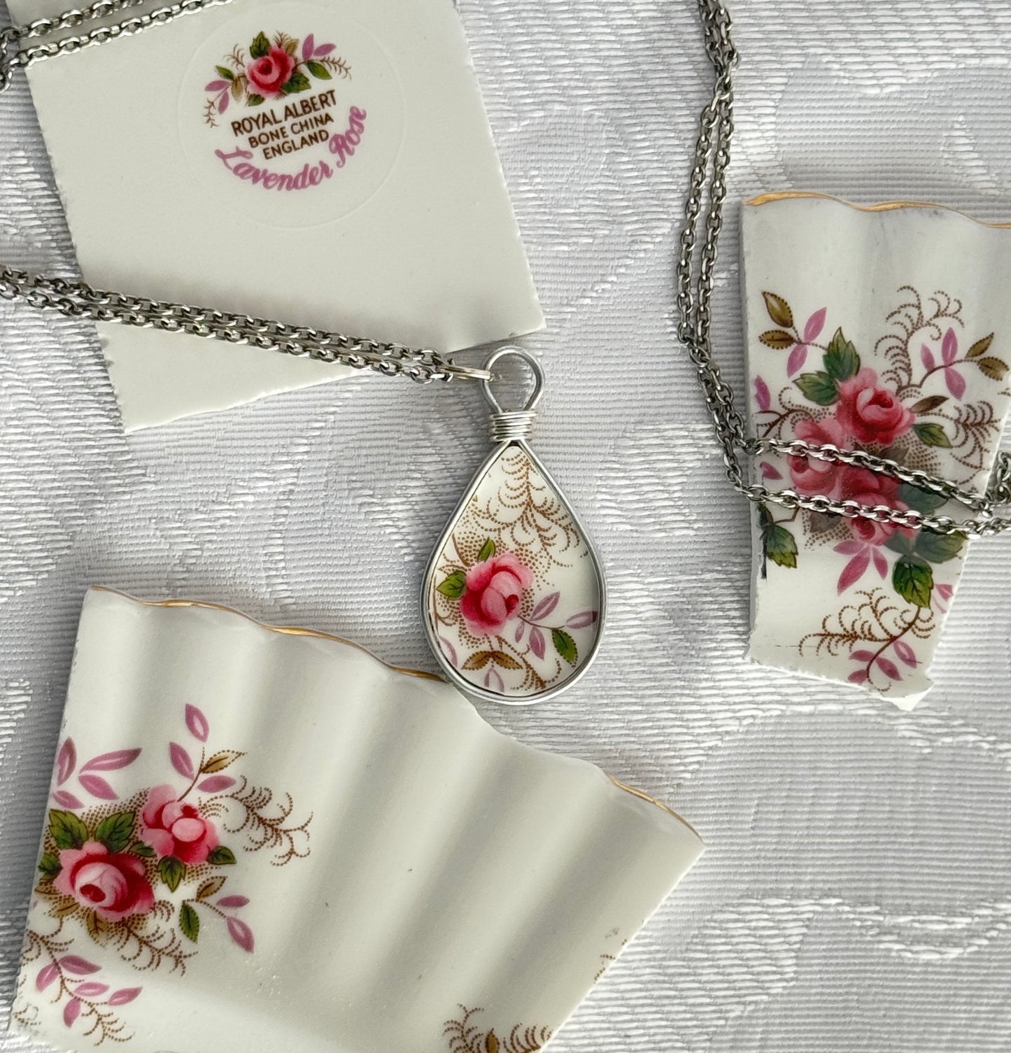 Teardrop-shaped pendant with floral design on a chain, surrounded by matching fabric and card on a textured surface.