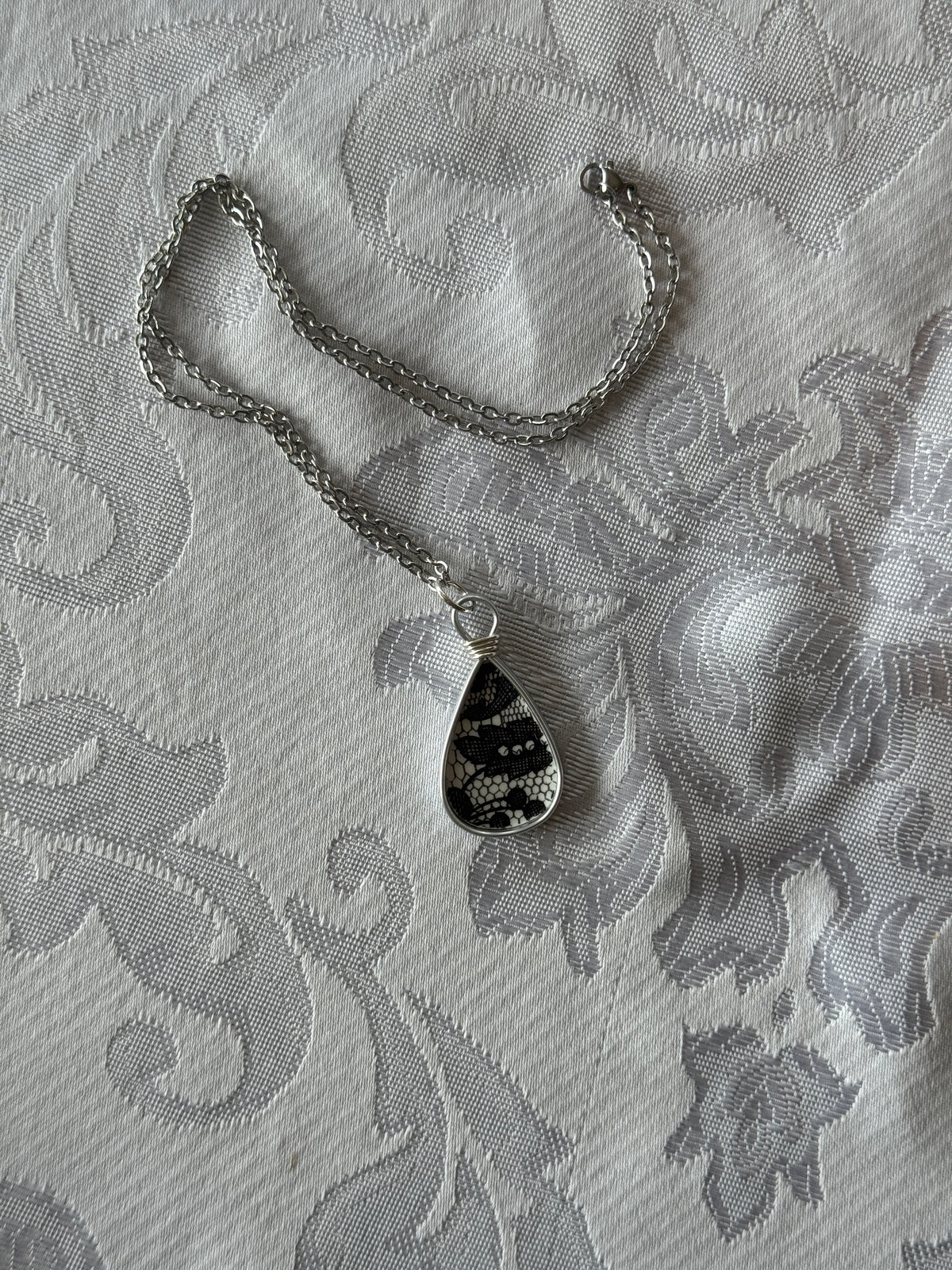 A silver chain necklace with a teardrop-shaped pendant featuring a black and white snakeskin pattern, laid flat on a white patterned fabric background.