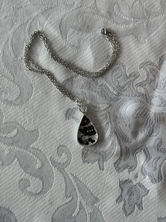 A silver chain necklace with a teardrop-shaped pendant featuring a black and white snakeskin pattern, laid flat on a white patterned fabric background.