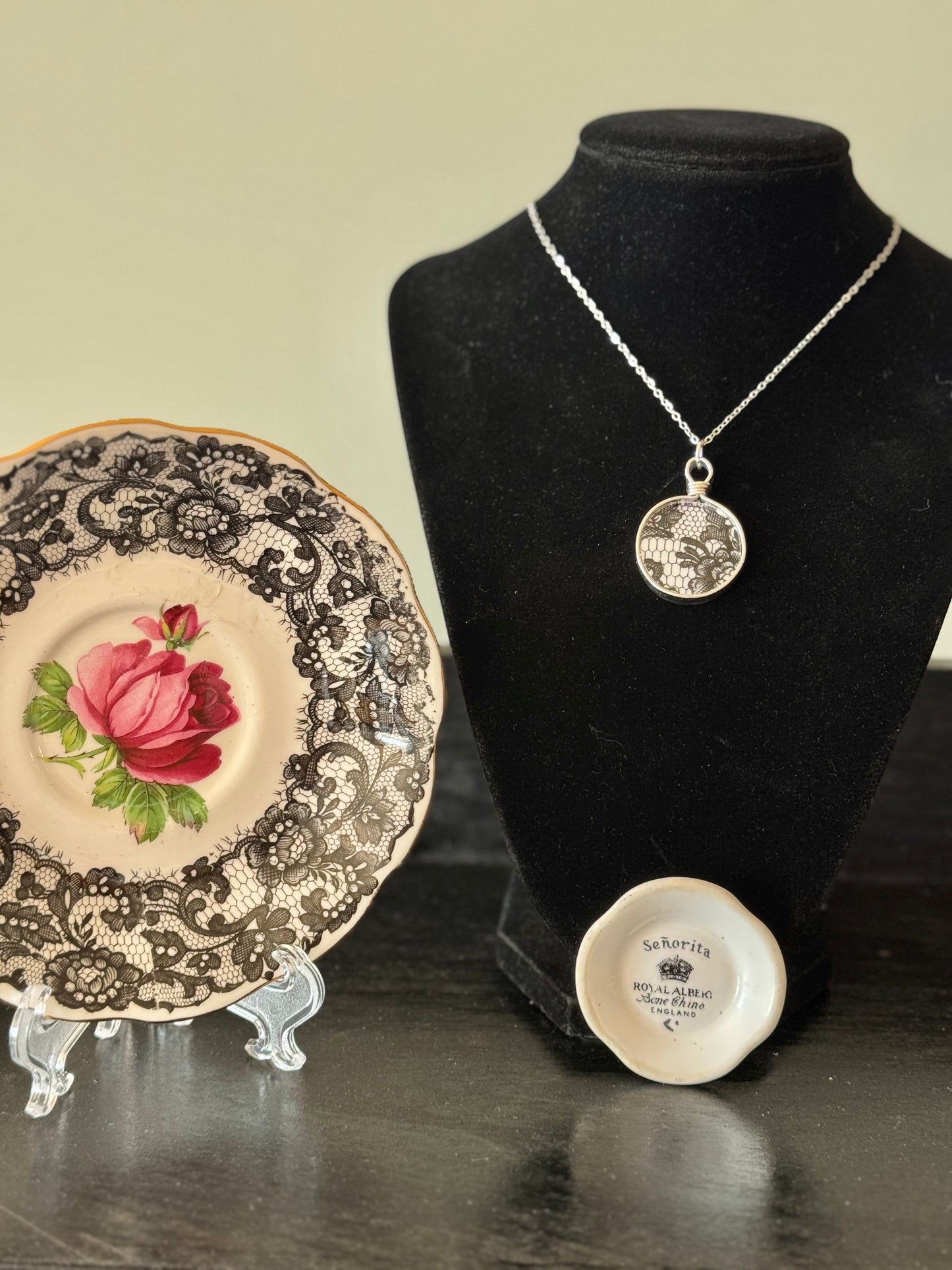 Necklace displayed on a black mannequin bust with a decorative plate and small round object on a reflective surface.