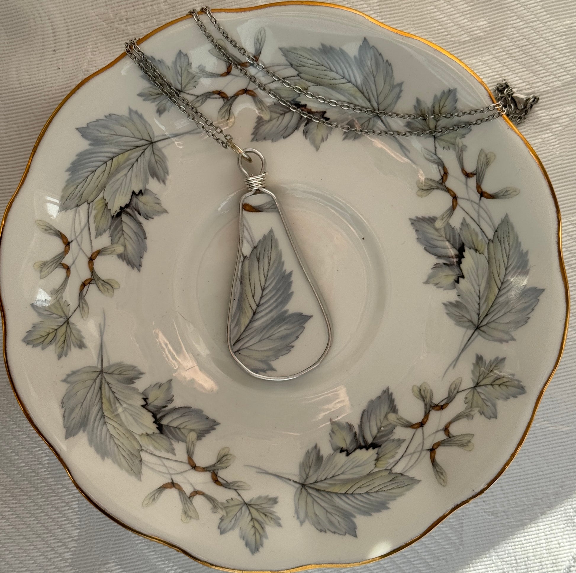 Teardrop-shaped pendant on a chain displayed on a decorative plate with leaf patterns.