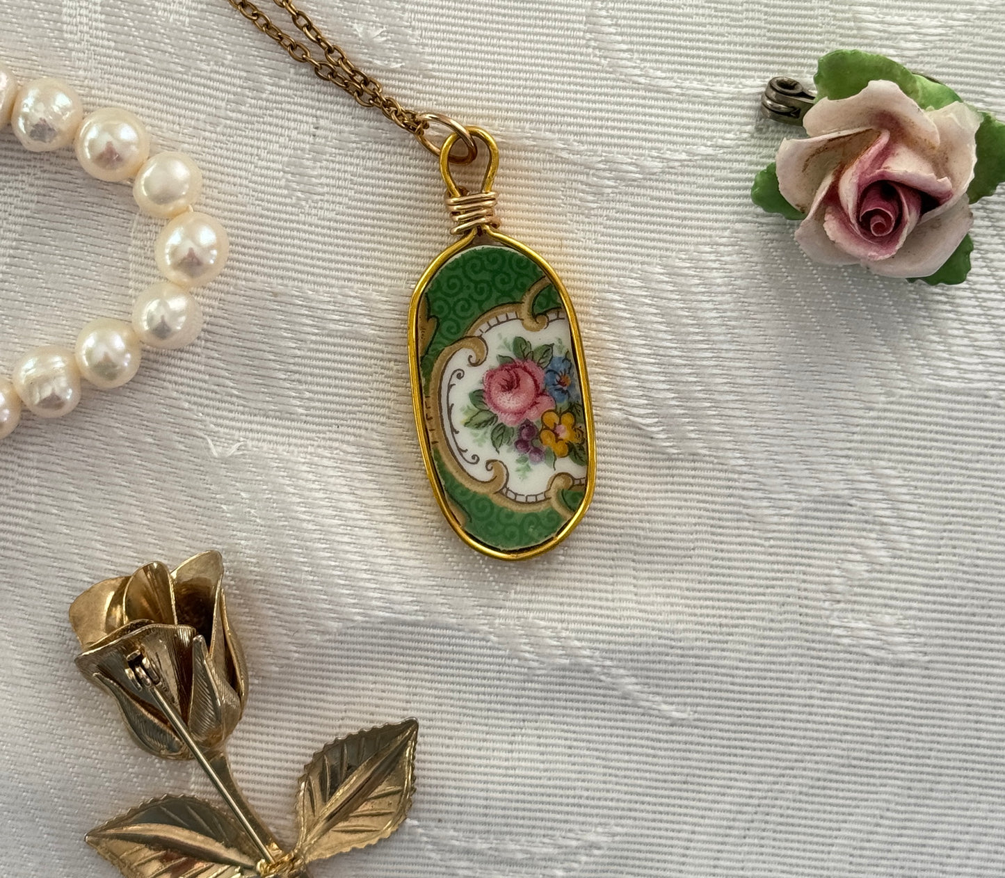 Vintage-style necklace with floral pendant, pearl bracelet, and gold brooch on a textured white surface.