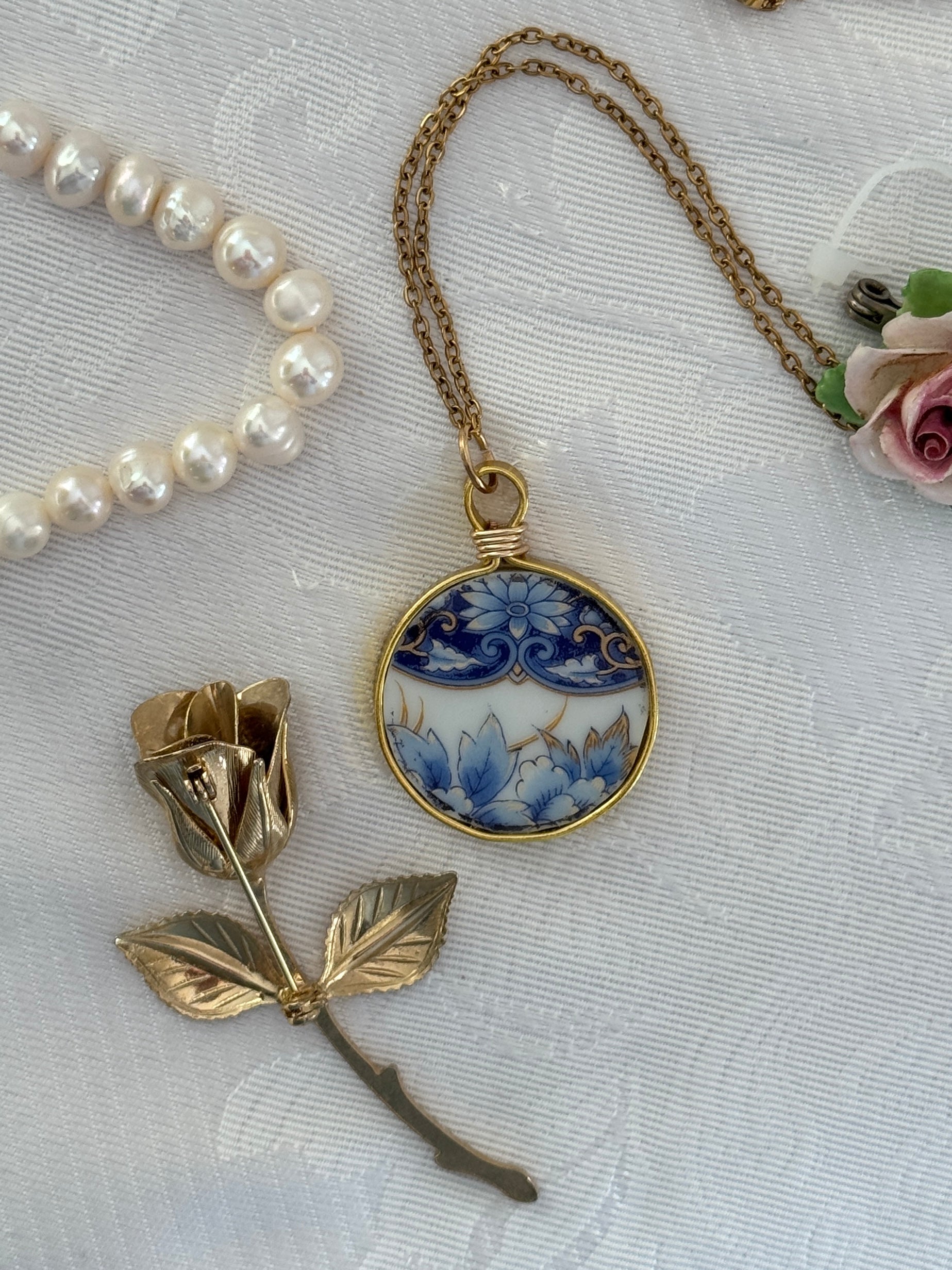 Necklace with a blue floral pendant and gold rose brooch on a textured white surface