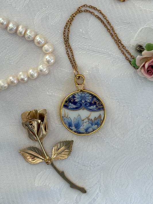 Necklace with a blue floral pendant and gold rose brooch on a textured white surface