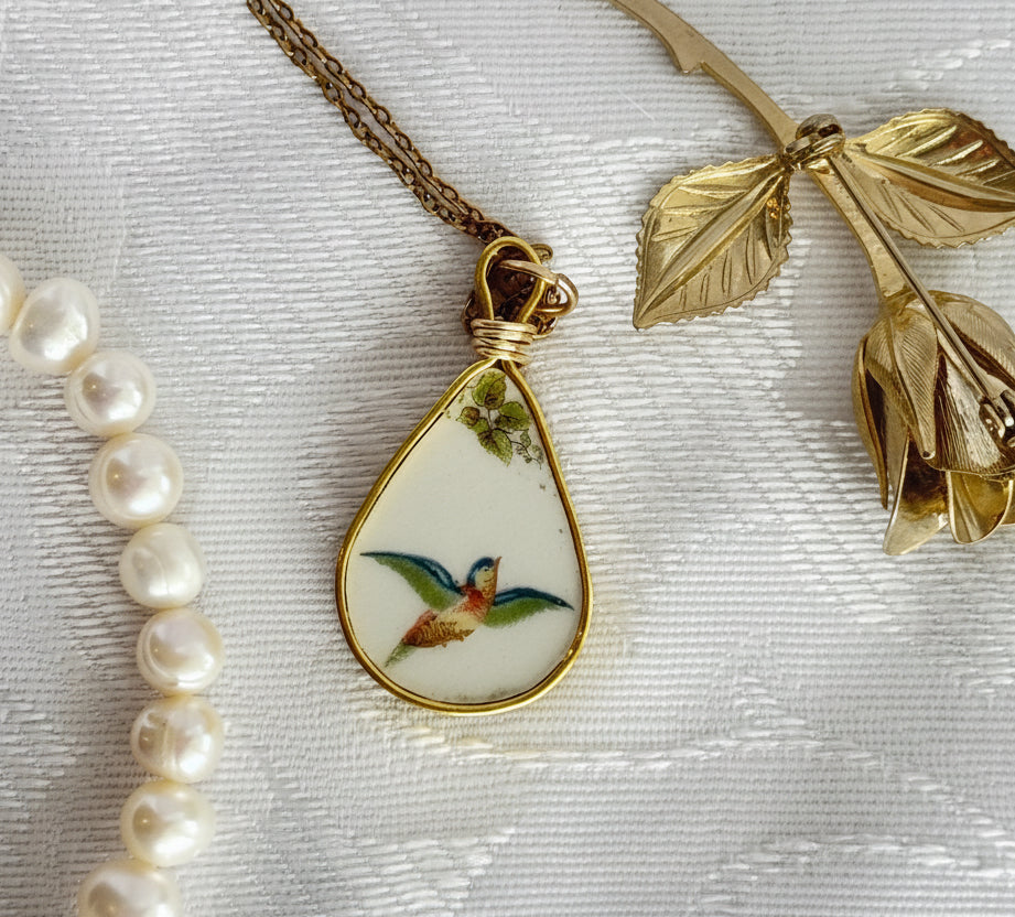 Gold necklace with a teardrop pendant featuring a bird design, pearl bracelet, and gold brooch on a textured white surface.